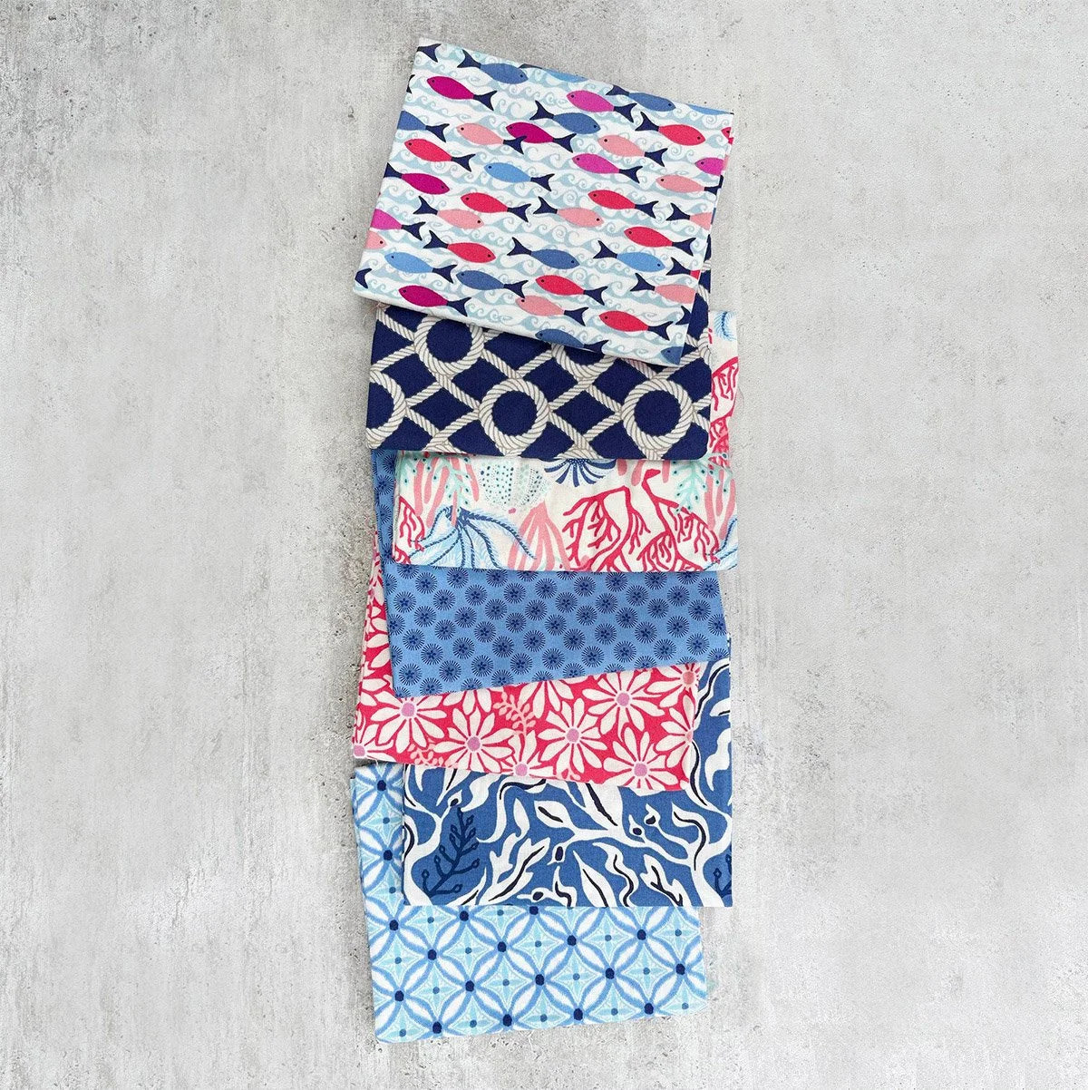 A vertical stack of nine folded fabric squares with various blue, red, and white patterns, displayed on a textured gray concrete surface.