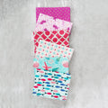 Five folded fabric napkins or small fabric squares stacked vertically on a textured gray concrete background. The fabrics feature various colorful patterns including pink polka dots, white floral motifs, red lattice, turquoise mermaids, and blue fish.