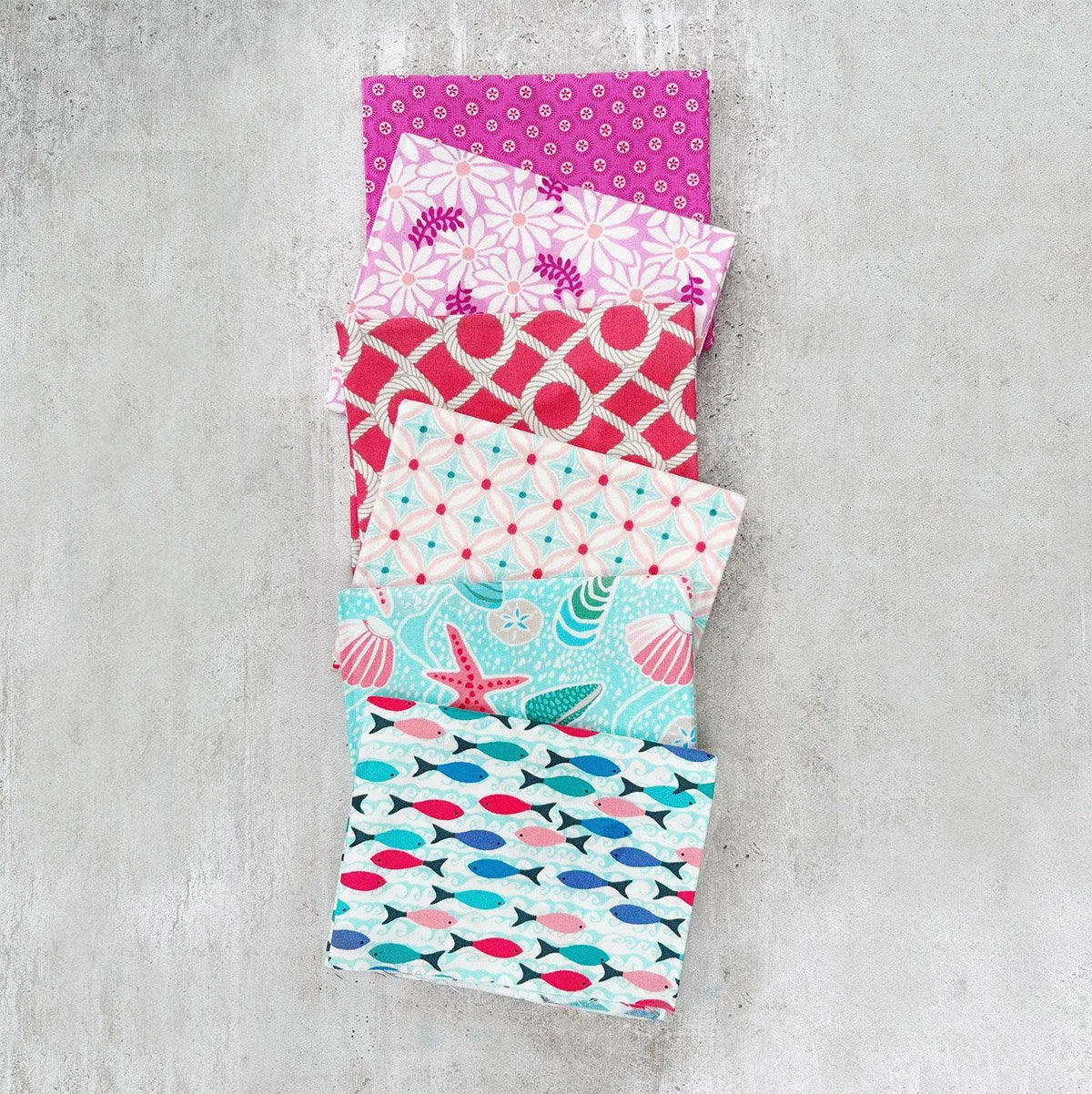 Five folded fabric napkins or small fabric squares stacked vertically on a textured gray concrete background. The fabrics feature various colorful patterns including pink polka dots, white floral motifs, red lattice, turquoise mermaids, and blue fish.