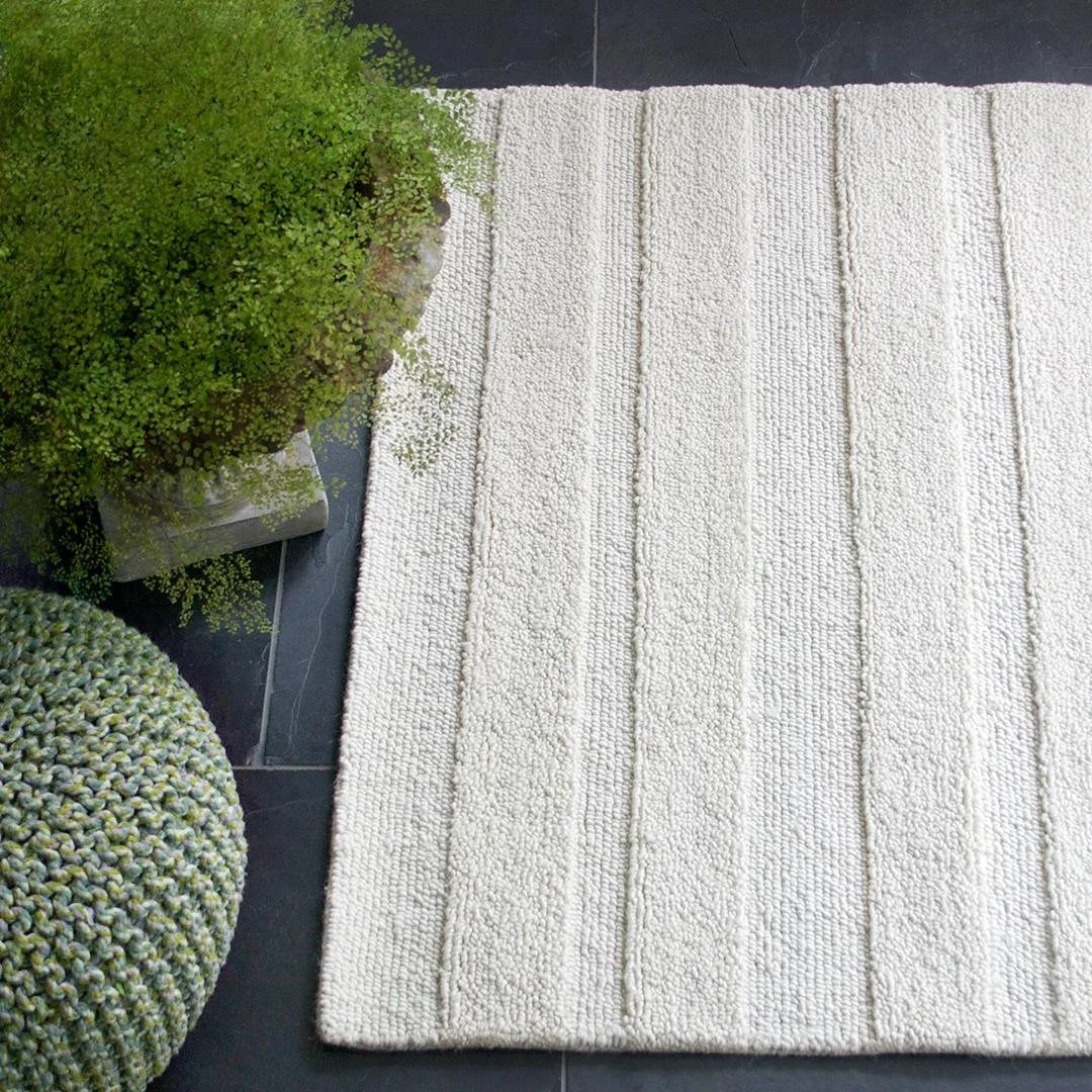 eye heart hand Pure Stripe hand-tufted rug in undyed natural white wool with subtle tonal stripes, on a slate floor beside a knit pouf and fern – Kate Spain design