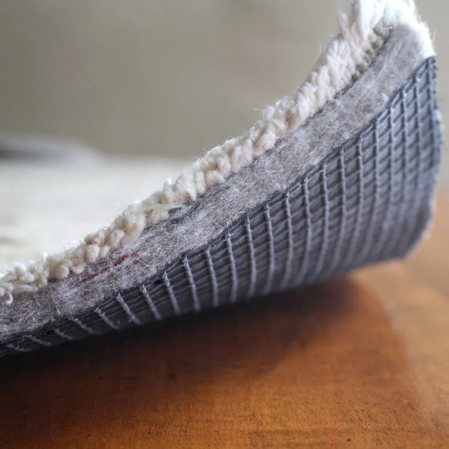 Premium rug pad of recycled felt with natural rubber grid backing, corner of a rug turned up to show the pad's rubber grid