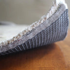 Premium rug pad of recycled felt with natural rubber grid backing, corner of a rug turned up to show the pad's rubber grid
