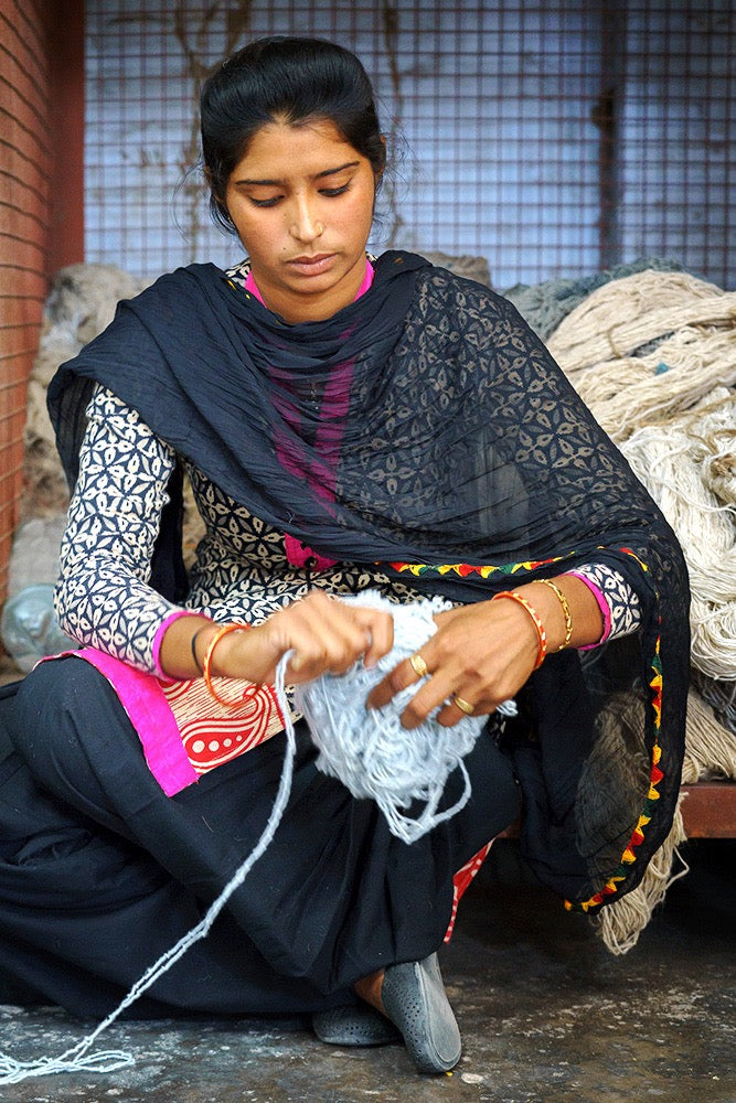 Artisan in India seated among wool bundles, winding pale blue yarn by hand