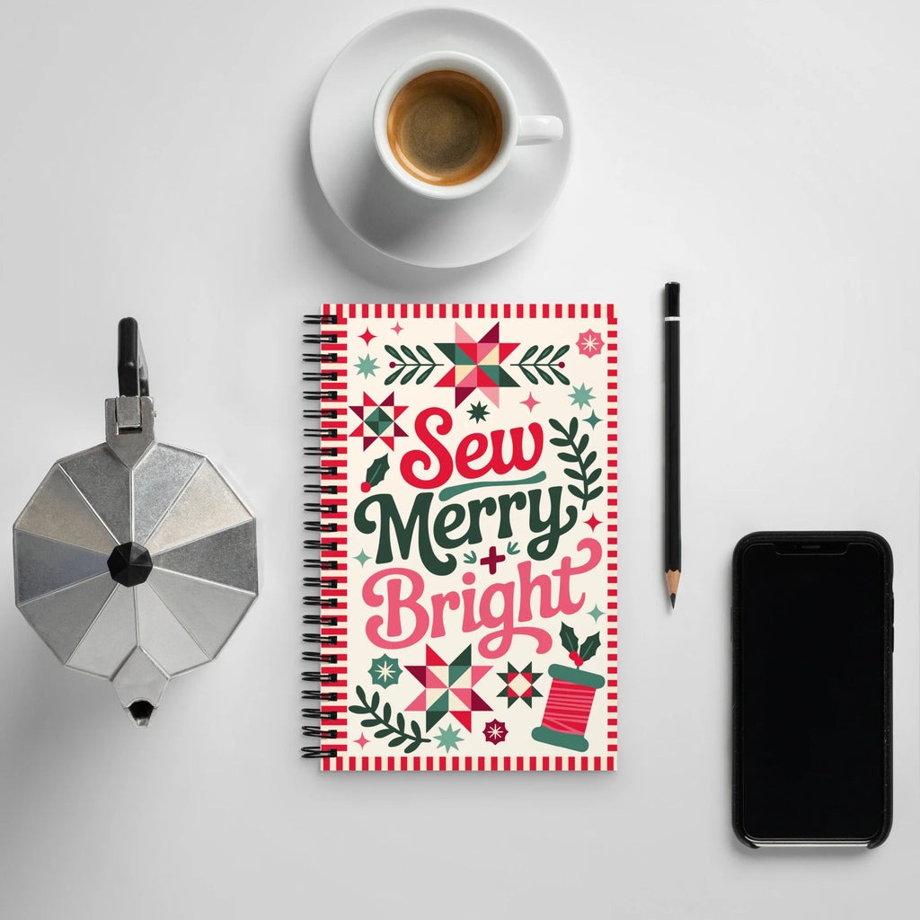 Sew Merry + Bright spiral notebook with quilt-block stars and holly, on a desk with coffee – Kate Spain design for Pat Sloan