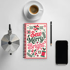Sew Merry + Bright spiral notebook with quilt-block stars and holly, on a desk with coffee – Kate Spain design for Pat Sloan