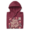 Sew Merry + Bright lettering with quilt-block stars, holly and a thread spool on a pullover hoodie in maroon, folded close-up of the design – Kate Spain design for Pat Sloan